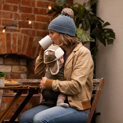 Mother outdoors holding a toddler’s hand while carrying a baby in a forest green wrap.