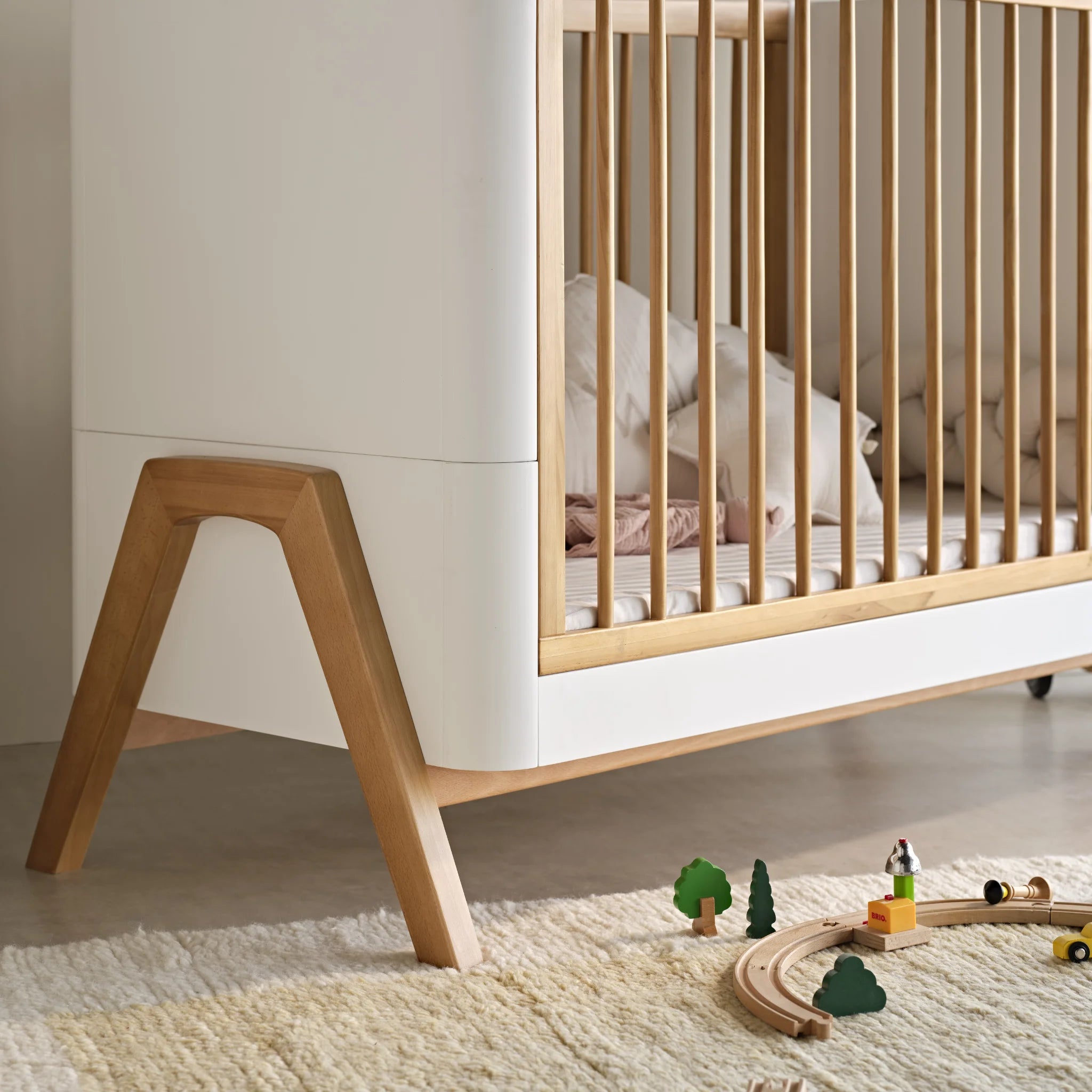 Close-up lifestyle-detail van het Gaia Baby Hera-ledikantbed met het ontwerp van de schuine poten met natuurlijk eikeneffect, de zijkanten van massief hout en de zachte, neutrale kinderkamerstijl met houten speelgoed en een vloerkleed met textuur.