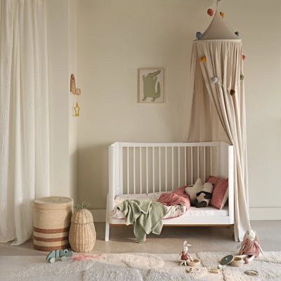Lifestyle-Bild des in ein Kleinkinderbett umgewandelten Gaia Baby Hera-Kinderbetts in Weiß, ausgestattet mit weicher Bettwäsche, Kissen und einem Stoffhimmel, eingerichtet in einem warmen, neutralen Kinderzimmer mit Spielzeug und gewebten Accessoires.