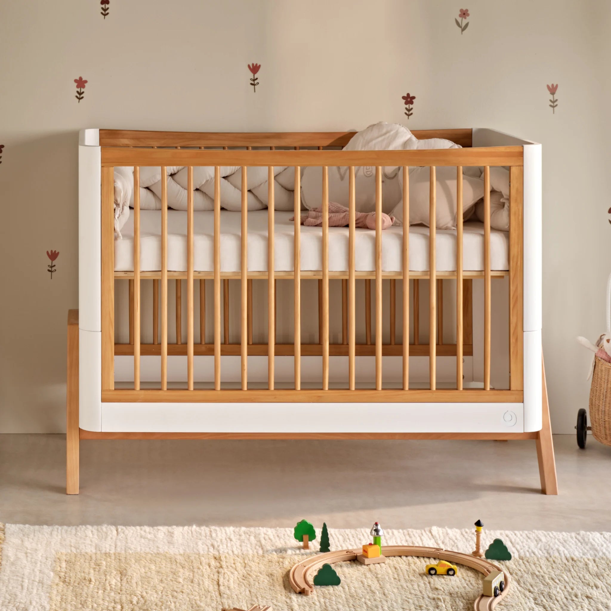 Hölzernes Gaia Baby Hera Kinderbett mit weißen Seitenwänden und einer weißen Matratze, gestaltet in einer warmen Kinderzimmerumgebung mit einer weich gemusterten Wand und auf einem Teppich im Vordergrund angeordneten Spielzeugen.