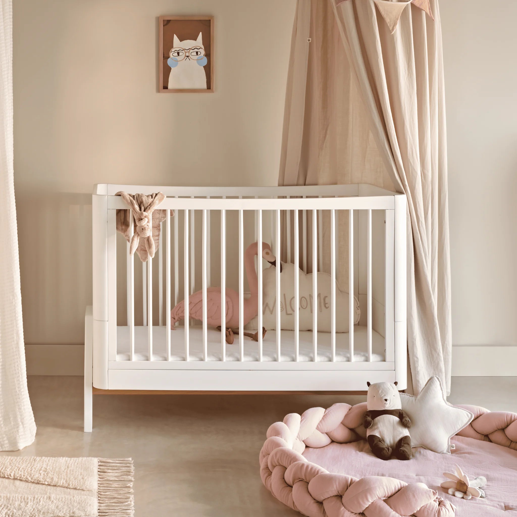 Lifestyle-afbeelding van het Gaia Baby Hera-ledikantbed in een geheel witte afwerking, gestyled in een rustige, neutrale kinderkamer met zachtroze accenten, een gedrapeerde hemel, knuffels en gevlochten vloerkussens.