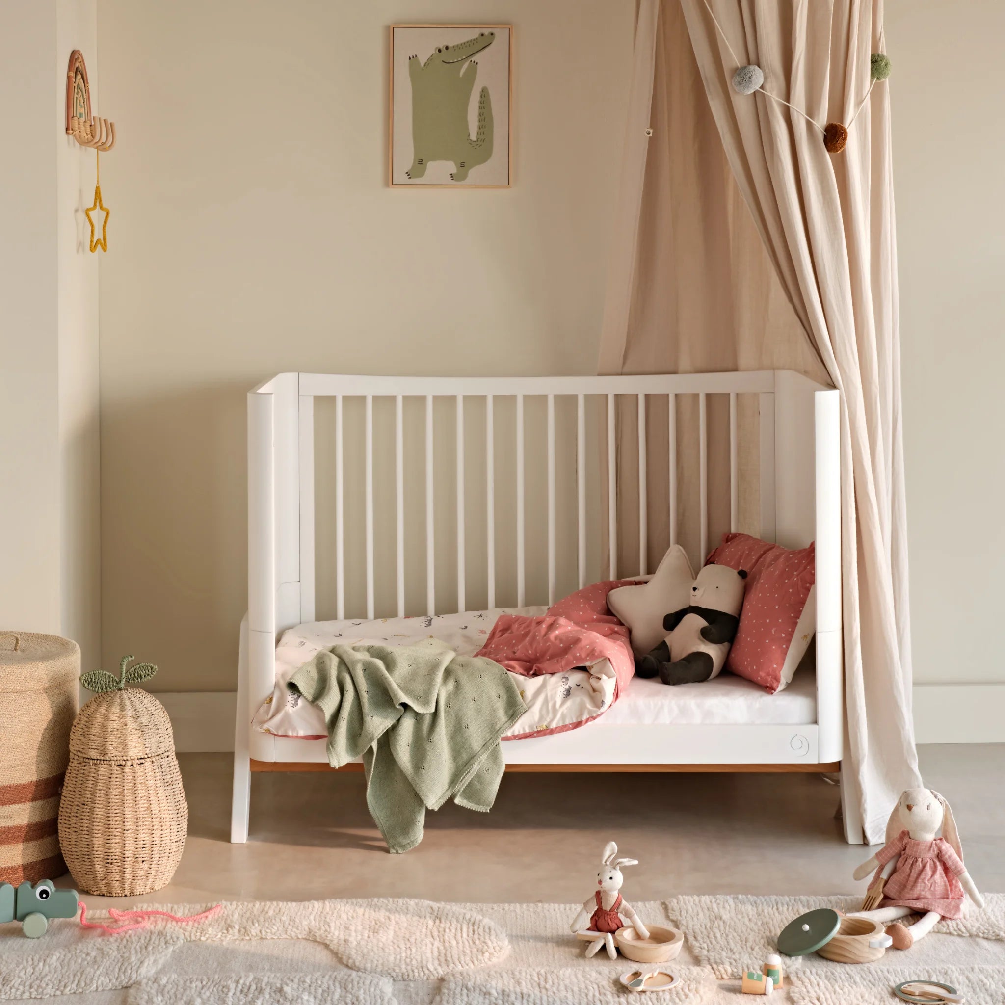 Lifestyle-afbeelding van het Gaia Baby Hera peuterbed in een geheel witte afwerking, gestyled in een zachte, neutrale kinderkameromgeving met een gedrapeerde hemel, pastelkleurig beddengoed, knuffels en geweven opbergmanden.