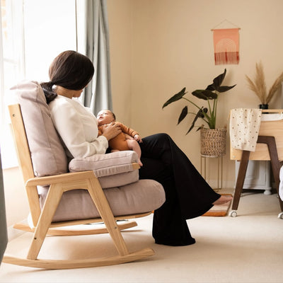 Gaia Baby Hera Rocking Chair in natural oak with blush pink cushions, modern wooden nursery glider chair for breastfeeding and newborn bonding, mother holding baby in Scandinavian style nursery room.