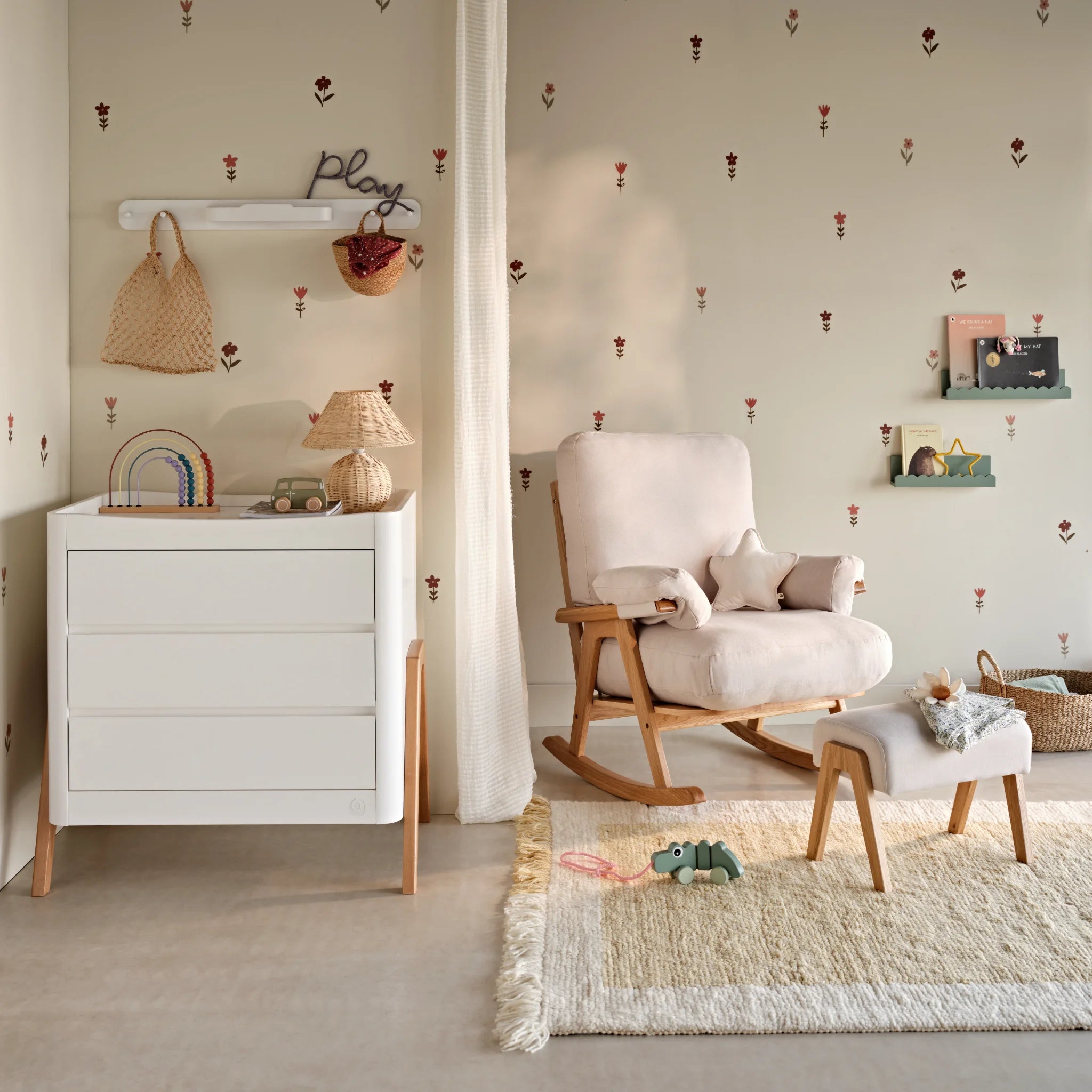 Styled nursery corner featuring a white Gaia Baby Hera dresser with natural wooden legs, paired with a cushioned rocking chair and footstool, surrounded by soft décor, wall hooks, and children’s toys on a rug.