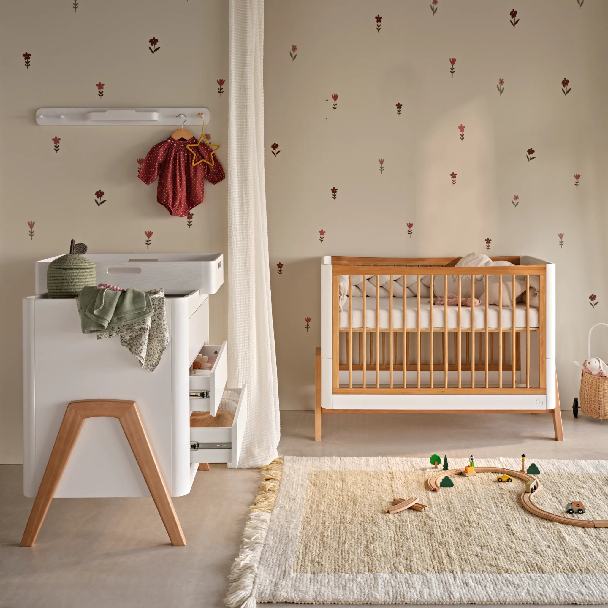 Kinderzimmerszene mit einer Gaia Baby Hera Kommode mit integrierter Wickelstation und Eichenbeinen neben einem passenden Holzbett. Der Raum liegt vor einer weich gemusterten Wand und verfügt über eine Kleiderstange mit Babykleidung, einen strukturierten Teppich mit Spielzeug und eine warme, neutrale Einrichtung.