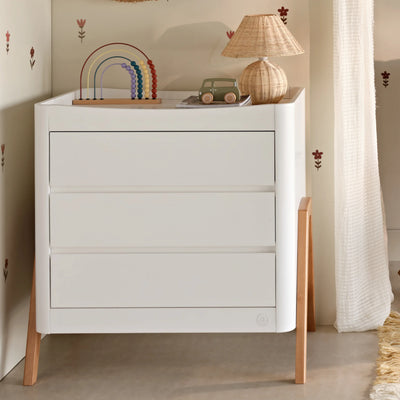 White Gaia Baby Hera dresser with natural wooden legs styled in a nursery setting, featuring a lamp, toy car, and rainbow stacking toy on top, against a softly decorated wall.