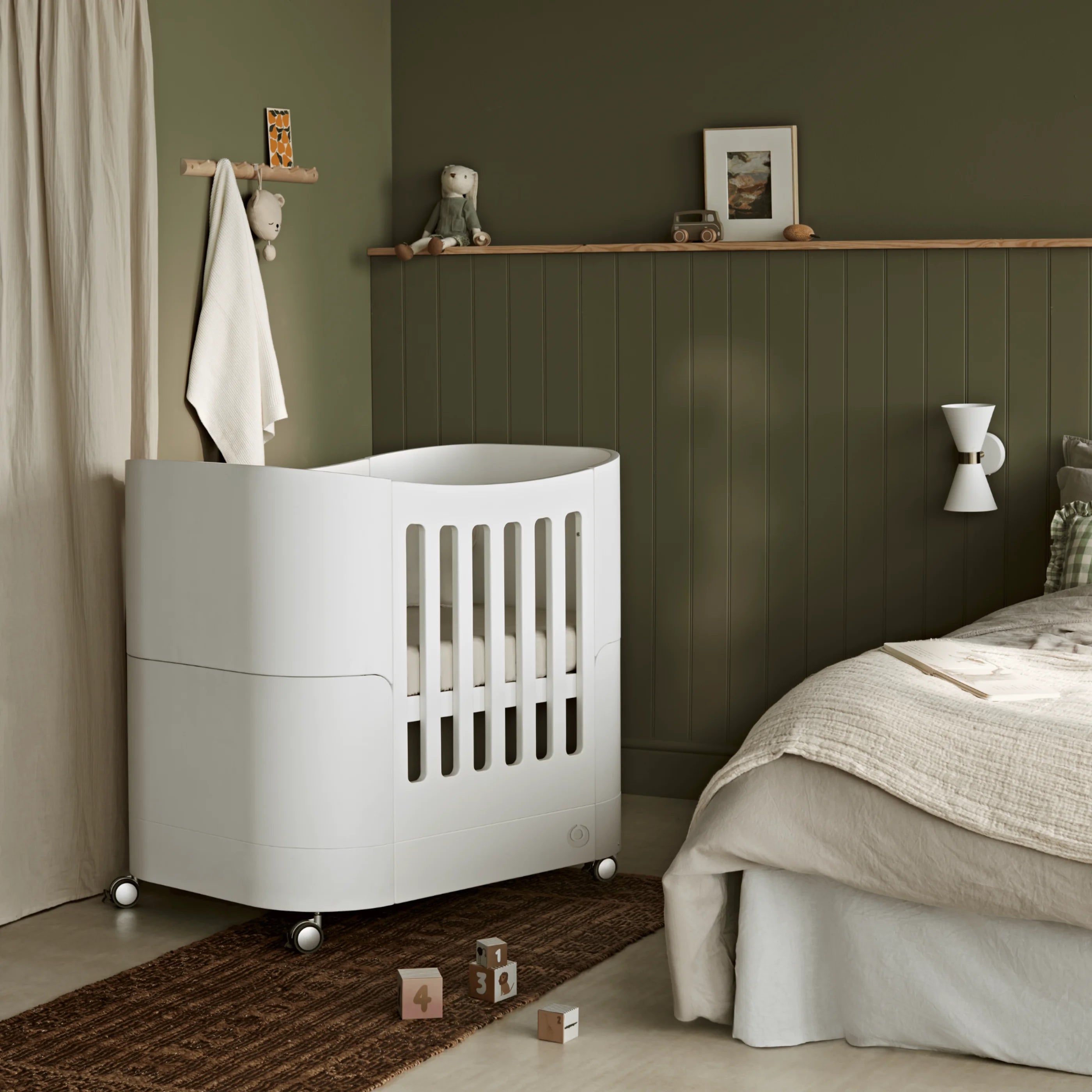 Gaia Baby Serena Mini-Kinderbett mit Rädern in Weiß, gestaltetes Kinderzimmer mit grün getäfelten Wänden und modernem, neutralem Dekor.