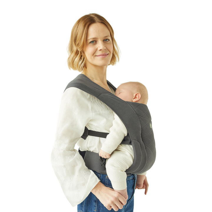 Blond haired mother wearing Gaia Baby Newborn Plus Carrier in Graphite. Sleeping newborn in a baby carrier