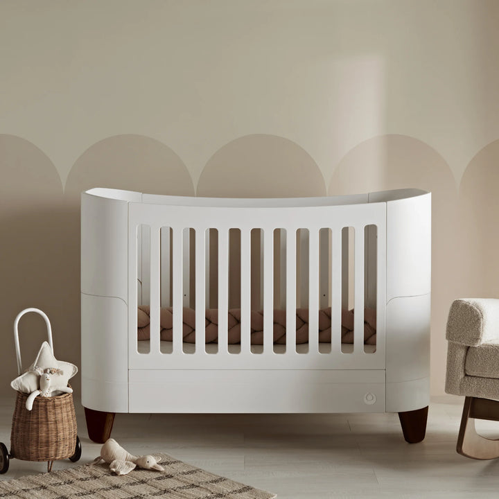 Elegant neutral nursery featuring the Gaia Baby Serena cot bed with walnut legs, styled with soft blush bedding, woven storage basket, textured rug, and warm beige wall detailing for a calm Scandinavian-inspired aesthetic.