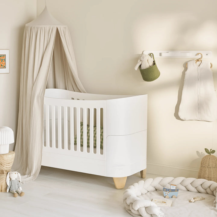 Modern neutral nursery featuring the Gaia Baby Serena cot bed with natural wood legs, styled with a soft beige canopy, braided floor cushion, hanging baby essentials, and Scandinavian-inspired décor in warm natural tones.